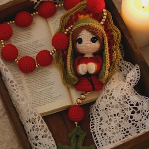 Puede incluir: Un rosario hecho a mano con cuentas rojas y una cruz verde. La pieza central presenta una figura de mu&ntilde;eca con una t&uacute;nica roja y un chal verde. El rosario se exhibe en una bandeja de madera con un libro y una vela encendida.