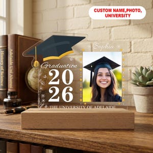 May include: A graduation photo frame with a wooden base. The clear acrylic frame features a graduation cap graphic, the year "2026", and the text "THE UNIVERSITY OF ADELAIDE". A photo of a smiling person is displayed.