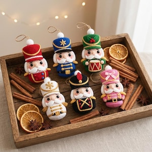 May include: Six crocheted nutcracker ornaments in red, blue, green, white, black, and pink. Each figure features a tall hat, a white moustache, and a hanging loop. The ornaments are arranged with dried orange slices, cinnamon sticks, and star anise.