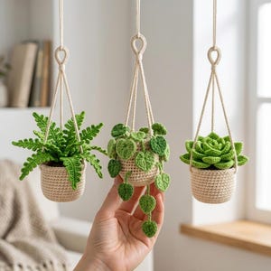 May include: Three hanging crochet plant decorations in beige pots. The left features a bright green fern, the center a trailing vine with heart-shaped leaves, and the right a succulent. Each is suspended by a cream-colored cord loop.
