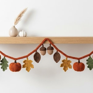 May include: A crochet garland featuring orange pumpkins, brown acorns, and various autumn leaves in shades of green, yellow, orange, and brown. The decorative piece is strung on a thick, braided orange yarn cord, creating a festive seasonal display.