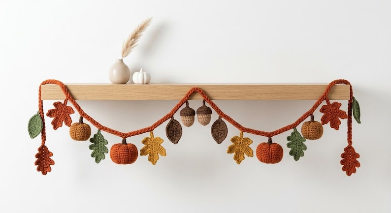 May include: A crochet garland featuring orange pumpkins, brown acorns, and various autumn leaves in shades of green, yellow, orange, and brown. The decorative piece is strung on a thick, braided orange yarn cord, creating a festive seasonal display.