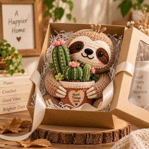Puede incluir: Un juguete de perezoso tejido a ganchillo con pelaje marrón y manchas oscuras en los ojos, que sostiene una maceta color terracota con tres cactus verdes y flores rosas. La maceta tiene una etiqueta en forma de corazón con el texto Good Things Grow.