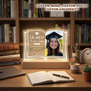 May include: A graduation gift in the shape of an open book, illuminated with a photo and text. The text includes "She did it!", a name, university, and graduation year. The frame is set on a wooden base.