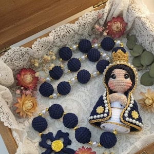 Puede incluir: Rosario de ganchillo hecho a mano con cuentas azul marino y una flor decorativa. El rosario incluye una mu&ntilde;eca de ganchillo de la Virgen Mar&iacute;a, adornada con una corona dorada y una capa azul y blanca. El rosario se exhibe en una caja de madera.