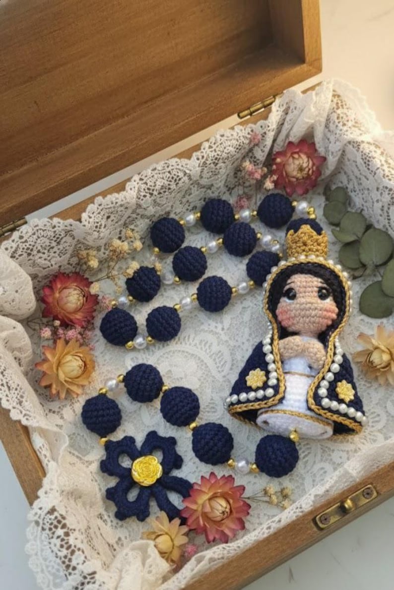 Puede incluir: Rosario de ganchillo hecho a mano con cuentas azul marino y una flor decorativa. El rosario incluye una mu&ntilde;eca de ganchillo de la Virgen Mar&iacute;a, adornada con una corona dorada y una capa azul y blanca. El rosario se exhibe en una caja de madera.