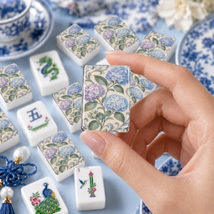 May include: A collection of rectangular mahjong tiles featuring various designs, including blue and purple hydrangea floral patterns, a green dragon, a blue peacock, a hummingbird, and the blue Chinese character for five on a white background.