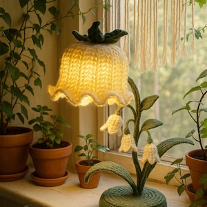 Könnte beinhalten: Handgefertigte, blütenförmige Lampe mit cremefarbenem Strickschirm und grünem Stiel und Sockel. Die Lampe hat drei weiße, glockenförmige Blüten und steht auf einer Fensterbank mit Topfpflanzen, was eine gemütliche, natürliche Ästhetik erzeugt.