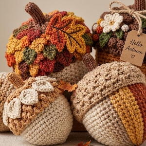 May include: A collection of crocheted acorn and pumpkin decorations in autumn colors. The items feature textured caps, leaves, and flowers. One acorn has a brown paper tag tied with twine that reads Hello Autumn with a heart symbol underneath.