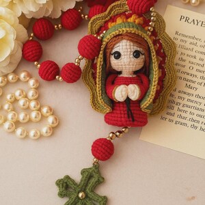 Puede incluir: Rosario de ganchillo hecho a mano con una mu&ntilde;eca vestida de rojo dentro de un marco dorado. El rosario incluye cuentas rojas, una cruz verde y detalles dorados. El fondo incluye flores y una tarjeta de oraci&oacute;n.