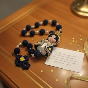 Puede incluir: Un rosario de ganchillo azul marino hecho a mano con detalles de perlas y una figura de ganchillo de la Virgen Mar&iacute;a. El rosario presenta una flor azul marino y una peque&ntilde;a tarjeta con texto escrito a mano. Confeti dorado esparcido alrededor.