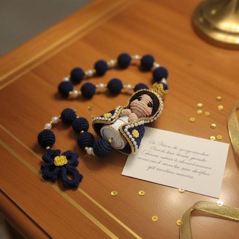 Puede incluir: Un rosario de ganchillo azul marino hecho a mano con detalles de perlas y una figura de ganchillo de la Virgen Mar&iacute;a. El rosario presenta una flor azul marino y una peque&ntilde;a tarjeta con texto escrito a mano. Confeti dorado esparcido alrededor.