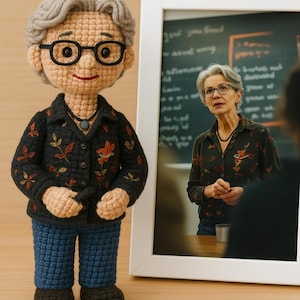 May include: A crocheted doll of a woman wearing glasses, a black jacket with orange and yellow floral embroidery, blue pants, and black shoes. A framed photo of the woman is next to the doll. The doll has gray hair and a necklace.