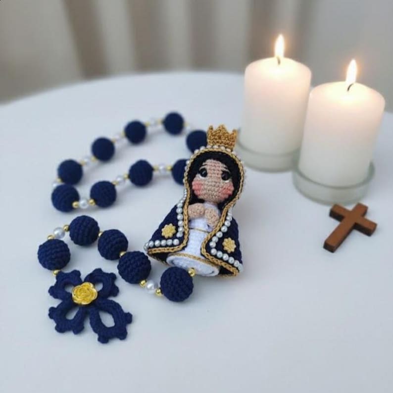 Puede incluir: Un rosario de ganchillo hecho a mano con cuentas azul marino y una cruz decorativa. Hay una peque&ntilde;a figura de ganchillo de la Virgen Mar&iacute;a, junto con dos velas encendidas y una cruz de madera. El rosario tiene una flor dorada.