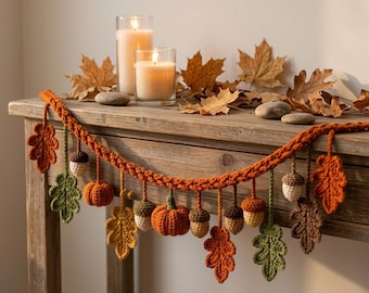 May include: A crocheted garland featuring orange pumpkins, brown and cream acorns, and various autumn-colored leaves in shades of green, yellow, and orange. The garland is draped across a wooden surface alongside two lit pillar candles.