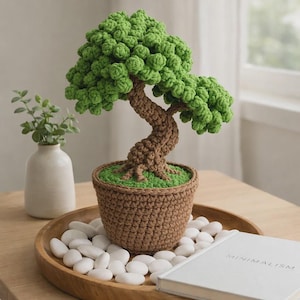 May include: A handcrafted crochet bonsai tree with a brown trunk and vibrant green foliage. The tree sits in a brown crochet pot with green 'grass' and is displayed on a wooden tray with white stones. A book with the word "MINIMALISM" is also on the tray.