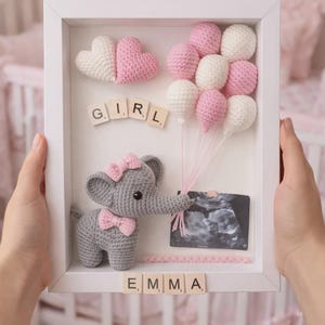 May include: A framed nursery decoration featuring a crocheted gray elephant with a pink bow, holding pink and white balloons. The frame includes the word "GIRL" and the name "EMMA" in letter tiles, along with a sonogram image. The background is a soft pink.