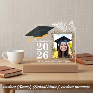 May include: A graduation photo frame with a black mortarboard design and the text "Graduation 2026 Let the Next Adventure Bloom." The frame includes a photo of a smiling person wearing a graduation cap, set on a wooden base.