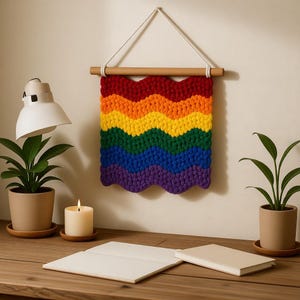 Puede incluir: Un tapiz de pared hecho a mano, de color arcoíris, con un diseño ondulado. La pieza está hecha de hilo grueso en tonos de rojo, naranja, amarillo, verde, azul y morado. Cuelga de una barra de madera y una cuerda blanca. El tapiz se exhibe en una pared beige.