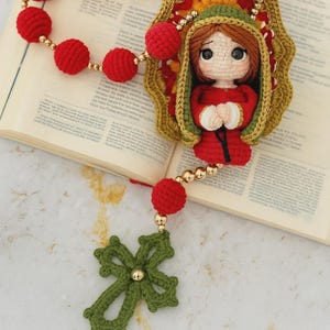 Puede incluir: Un rosario hecho a mano con ganchillo, con cuentas rojas y un colgante de cruz verde. La pieza central representa una figura de mu&ntilde;eca vestida de rojo y verde. Se incorporan cuentas y detalles dorados. El rosario se muestra sobre un libro abierto.