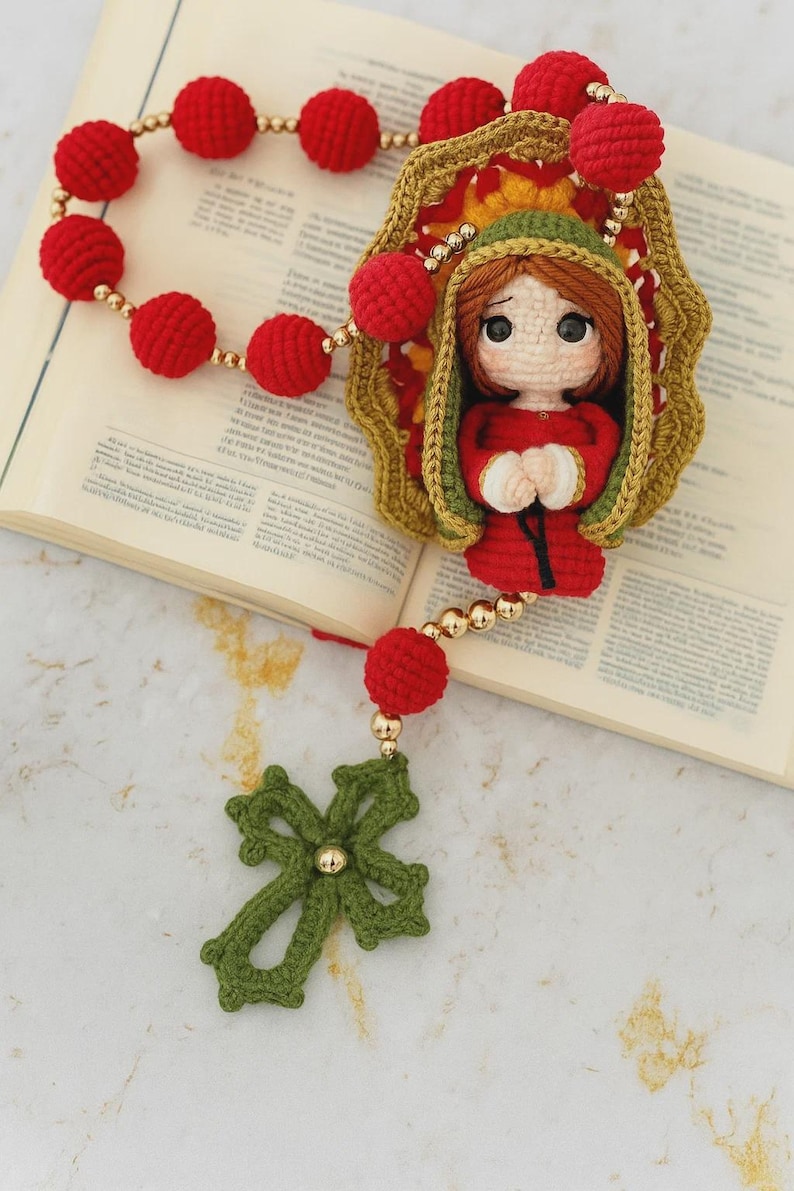 Puede incluir: Un rosario hecho a mano con ganchillo, con cuentas rojas y un colgante de cruz verde. La pieza central representa una figura de mu&ntilde;eca vestida de rojo y verde. Se incorporan cuentas y detalles dorados. El rosario se muestra sobre un libro abierto.
