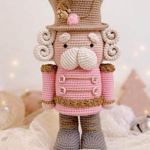 May include: A handmade crochet nutcracker doll in pink, tan, and gray. The doll has a tan top hat with floral accents, curly white hair, and a pink jacket with brown trim and buttons. Gray boots complete the festive design.
