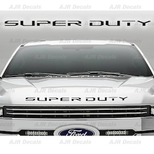 May include: A white Ford Super Duty pickup truck with a black "SUPER DUTY" decal on the hood and grille. The truck has a black Ford logo and headlights.