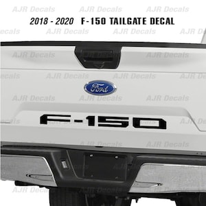 May include: A white Ford F-150 pickup truck tailgate with a black "F-150" decal. The truck is from the 2018-2020 model years.