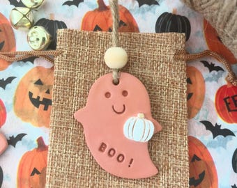 May include: A Halloween-themed craft project featuring a peach-colored ghost-shaped ornament holding a white pumpkin, with the word "BOO!" printed on it. The ornament hangs from a burlap bag. The background includes pumpkins, bats, and bells.