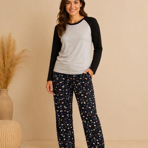 Puede incluir: Una camiseta de manga larga ragl&aacute;n con el torso gris jaspeado y mangas negras lisas, combinada con un pantal&oacute;n de pijama negro con un estampado repetitivo de peque&ntilde;os lazos y corazones coloridos en tonos rosa, azul y blanco por toda la tela.