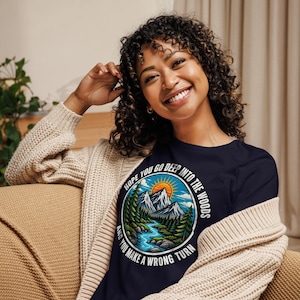May include: A dark navy blue t-shirt featuring a circular graphic of a mountain landscape with a river, pine trees, and a sun. The text surrounding the illustration reads, Hope you go deep into the woods and you make a wrong turn.