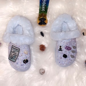 May include: A pair of light blue fuzzy slippers with rhinestones and various charms. One slipper has a tarot card patch and the other has a meditating figure charm.