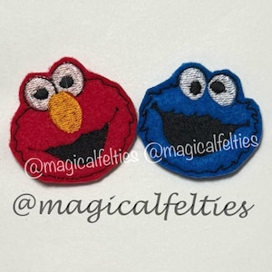 May include: Two embroidered felt patches featuring the faces of a red character with a yellow nose and a blue character with a black mouth. Both have white eyes with black pupils. The text @magicalfelties is visible on both patches and below.