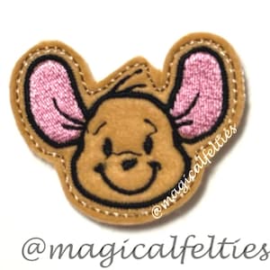 May include: A tan felt applique featuring an embroidered cartoon character head with large pink inner ears, black stitched facial features, and a smiling expression. The text @magicalfelties is embroidered in white on the right side and printed below.