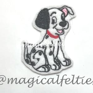 May include: A white felt Dalmatian puppy with black spots, a black ear, and a red collar. The puppy is sitting with its tail curled. The text "@magicalfelties" is written in cursive below the puppy.