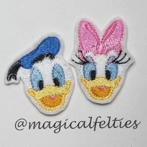 May include: Two embroidered felt appliques featuring cartoon character faces. One has a blue sailor hat and the other has a pink bow. Both have yellow beaks, blue eyes, and are outlined in white. The text "@magicalfelties" is at the bottom.