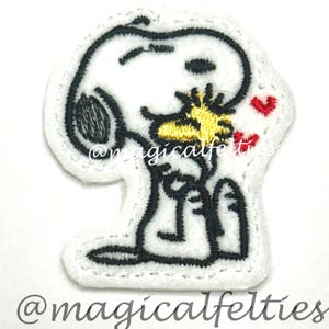 May include: A white felt patch featuring Snoopy kissing Woodstock, with red heart accents. The patch has black outline stitching and the text "@magicalfelties".
