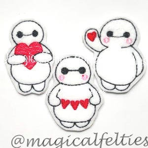 May include: Three white felt character appliques with black outlines. Each character has black eyes and a pink blush. One holds a red heart, another has a red heart, and the third has three red hearts. The text "@magicalfelties" is at the bottom.