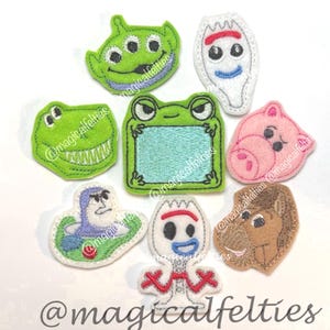 May include: A collection of eight embroidered felt patches featuring various cartoon character faces and shapes, including a green three-eyed alien, a white spork character, a green dinosaur, a green frog frame, a pink pig, a space ranger, and a brown horse.