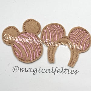May include: Two felt Mickey Mouse ear headbands. The ears are tan with pink striped detailing. The text "@magicalfelties" is printed on the ears. The ears are on a white background.