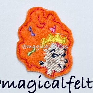 May include: An orange felt patch with an embroidered cartoon character wearing a yellow crown and glasses. The patch has a wavy outline and small embroidered details in pink, purple, and green. The text "@magicalfelties" is below the patch.