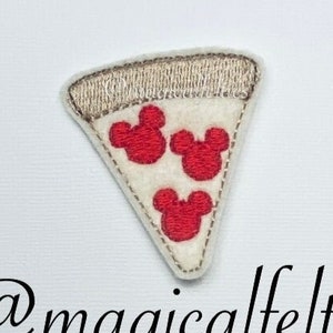 May include: A felt appliqué of a pizza slice with three red Mickey Mouse heads as toppings. The pizza slice is beige with a brown crust. The text "@magicalfelties" is below the pizza slice.