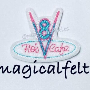 May include: A white felt patch with a pink and blue embroidered design of a stylized 'V' with the number '8' inside and the text 'Bob's Cafe' below. The patch is for sale by @magicalfelties.
