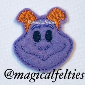 May include: Purple felt character with orange felt ears and black embroidered eyes. The character is smiling. The felt character is a handmade applique.
