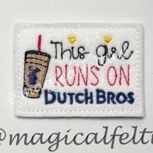 May include: A white felt patch with the embroidered phrase "This girl runs on Dutch Bros." A coffee cup with a red straw is on the left. The text is in black, red, and blue thread. Two small yellow hearts are above the word "girl".