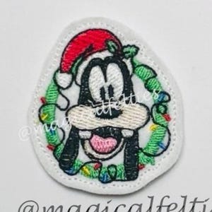 May include: Embroidered patch featuring Goofy in a Santa hat and a Christmas wreath. The patch has a white background and the text "@magicalfelties" at the bottom. The design includes red, green, black, and white colors.