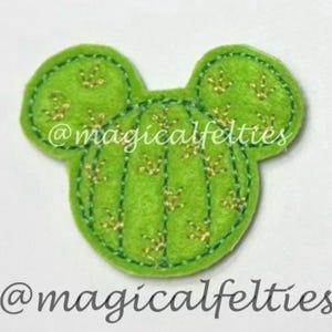 May include: A bright green felt applique shaped like a mouse head with a cactus-inspired design. The surface features vertical stitched lines and small gold-colored embroidered accents. The text @magicalfelties is visible on the object.