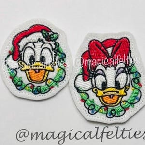May include: Two embroidered patches featuring cartoon duck characters in Christmas attire. One patch depicts a duck wearing a Santa hat and a wreath. The other patch shows a duck wearing a red bow and a wreath. The patches are white with colorful details.