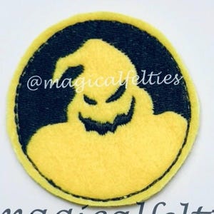May include: A round, yellow felt patch with a black border. The patch features a stylized, smiling ghost-like figure in black. The text "@magicalfelties" is printed above and below the patch.