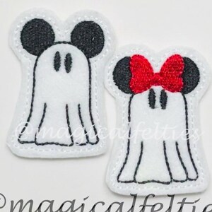 May include: Two felt ghost-shaped appliques. One has black mouse ears and a black face, the other has a red bow and black mouse ears. The appliques are white with black stitching. The text "@magicalfelties" is at the bottom.